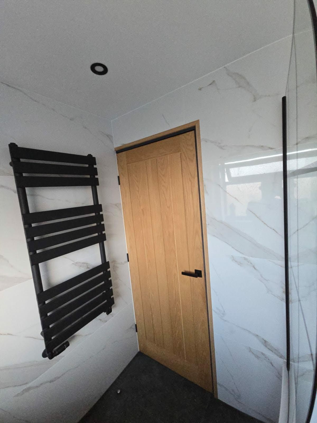 Oak door and black heated towel rail fitted against marble-effect bathroom tiling
